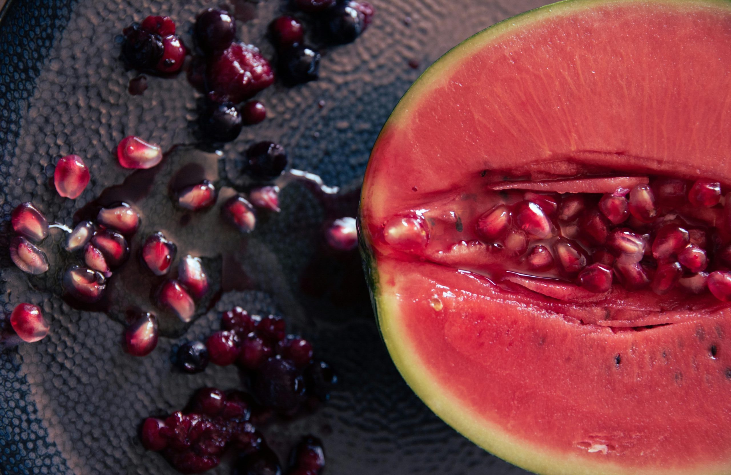 Sliced watermelon and pomegranate with seeds scattered on a dark surface highlighting fruit derived extracts and seeds used in natural skin care formulation