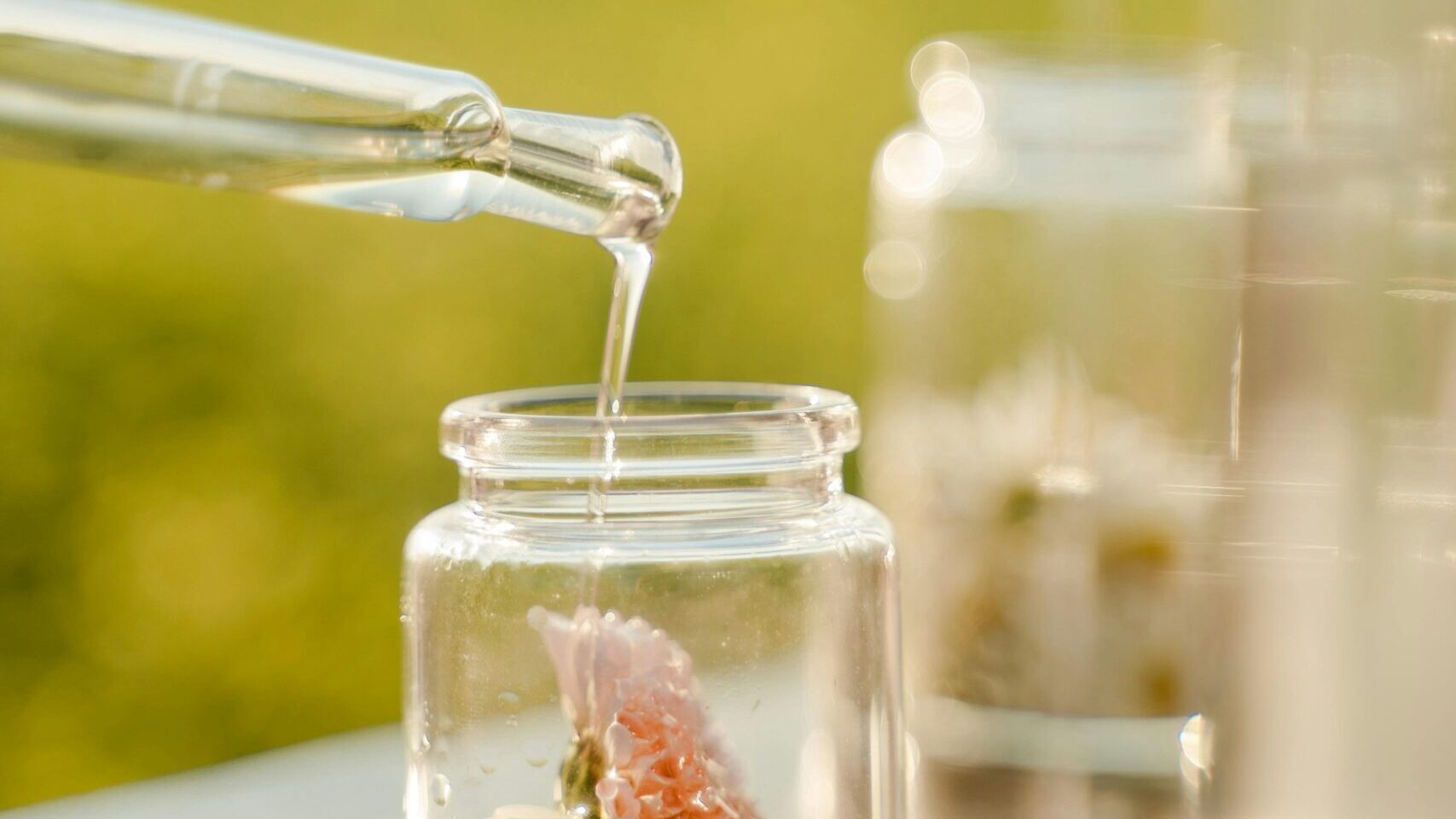 Glass dropper bottle pouring plant-based fragrance oil into a small jar with soft golden field in the background